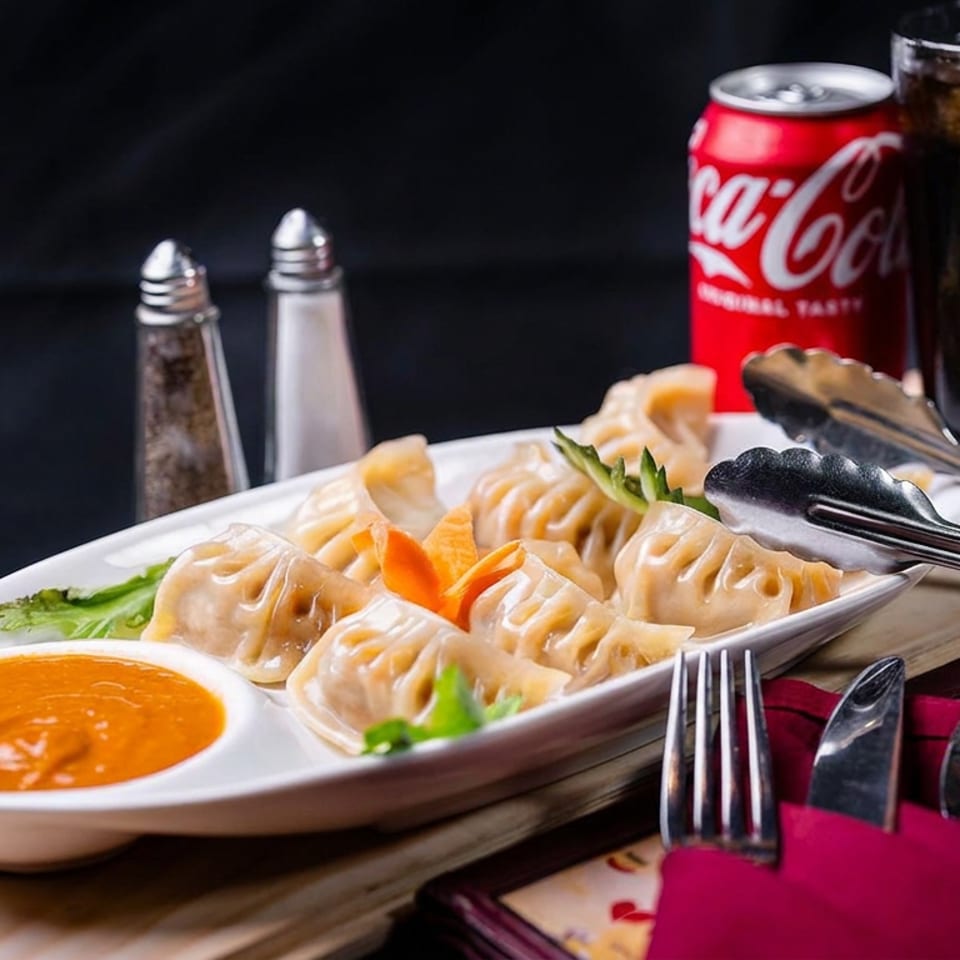 Steamed dumplings with orange dipping sauce, garnished with vegetables, alongside utensils, condiments, cola, and iced soda.